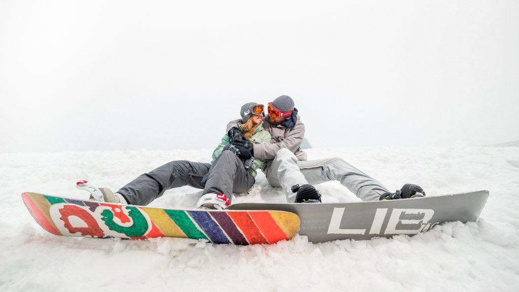 A couple at their snowshoe engagement photo shoot sitting down with snowboards.