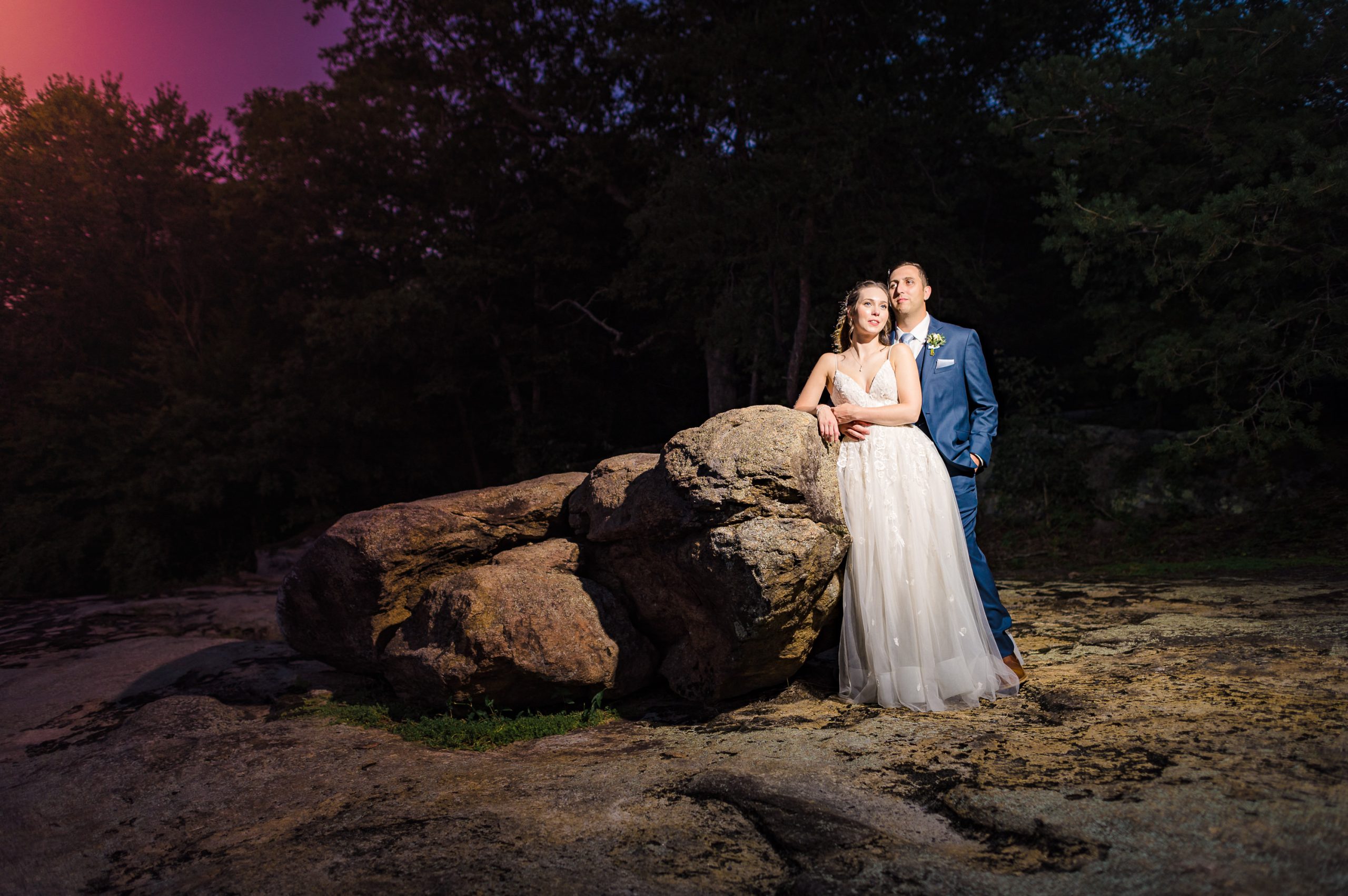 Bride and groom leaning on boulder darkly lit at twilight at the Mill at Fine Creek with off camera flash